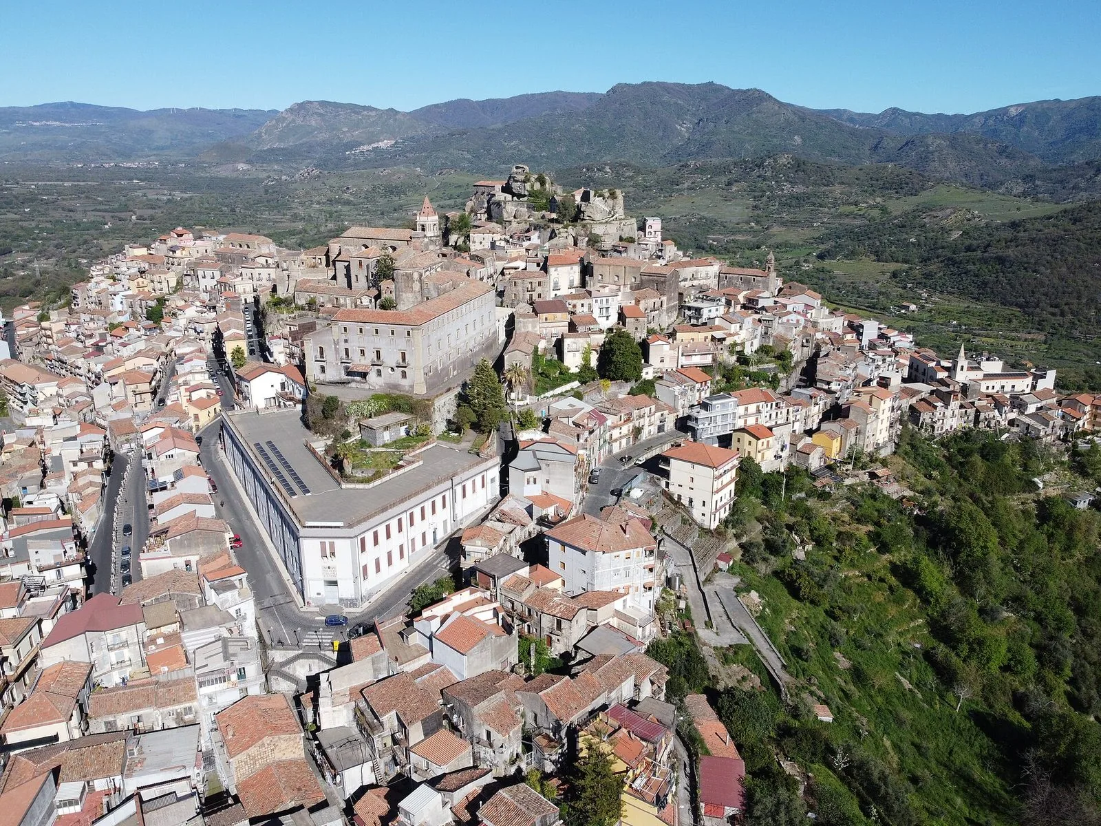 Veduta aerea di Castiglione di Sicilia: case dai tetti in coppi strette sul colle, la rupe del castello e i campanili in alto, con la valle verde e le montagne sullo sfondo sotto un cielo limpido