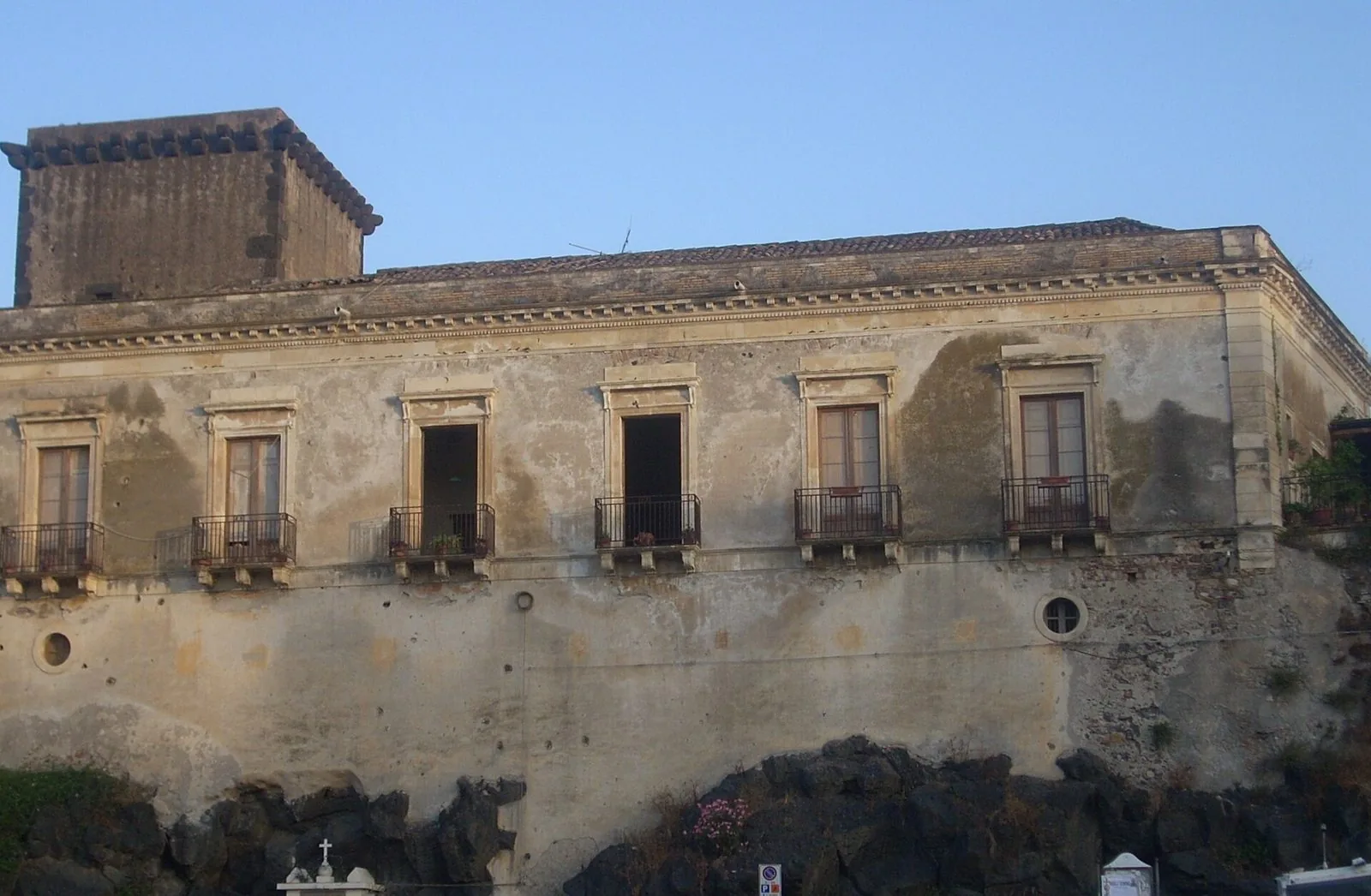 Facciata del Castello di Schisò con la torre squadrata in pietra scura, finestre con balconcini in ferro e la base appoggiata su scogli neri di lava, sotto un cielo azzurro