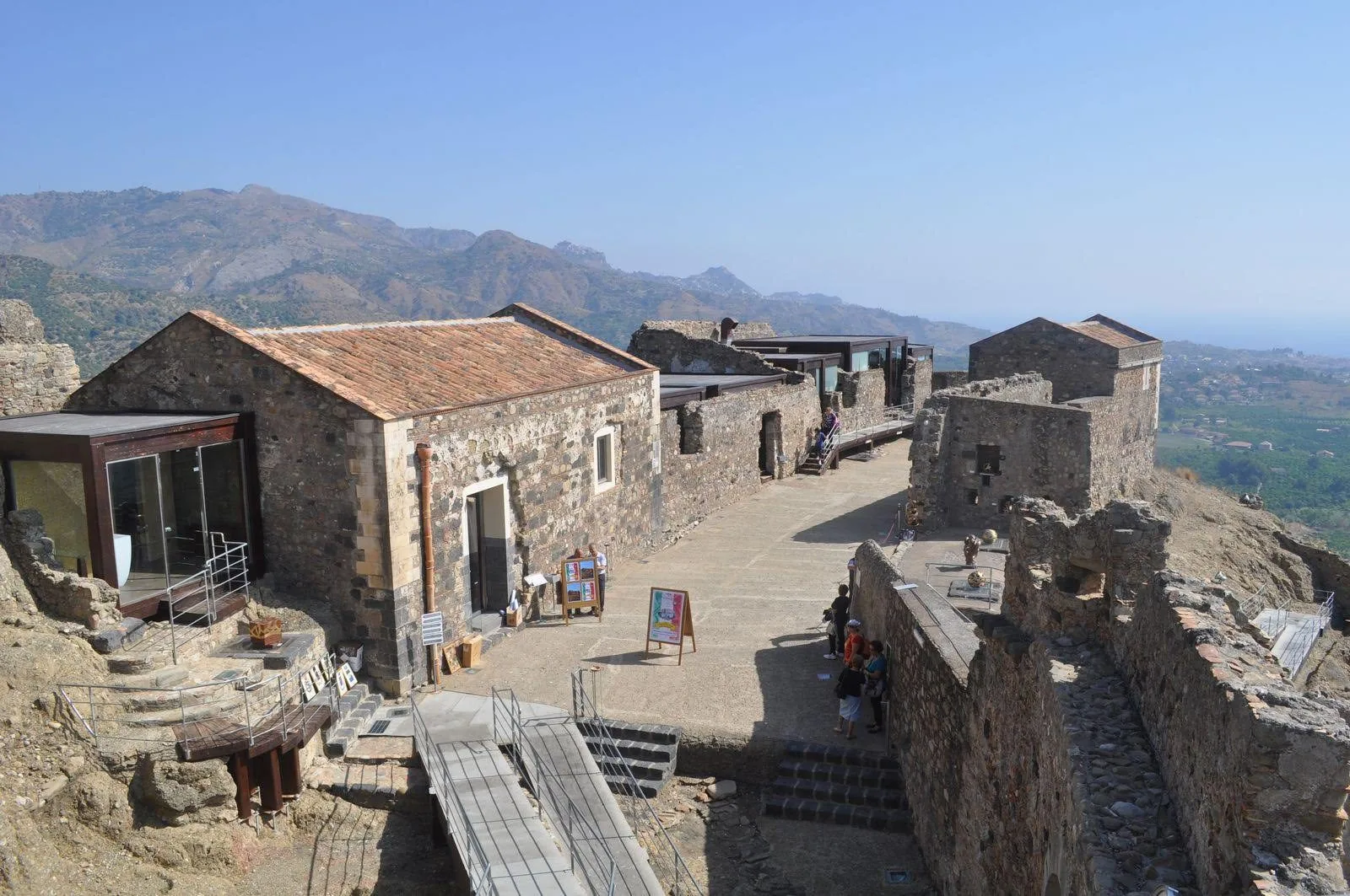 L'interno restaurato del Castello di Calatabiano: edifici in pietra lavica con tetti in coppi, una corte lastricata con passerelle e scale, alcuni visitatori in lontananza e, sullo sfondo, le colline verso Taormina e il mare