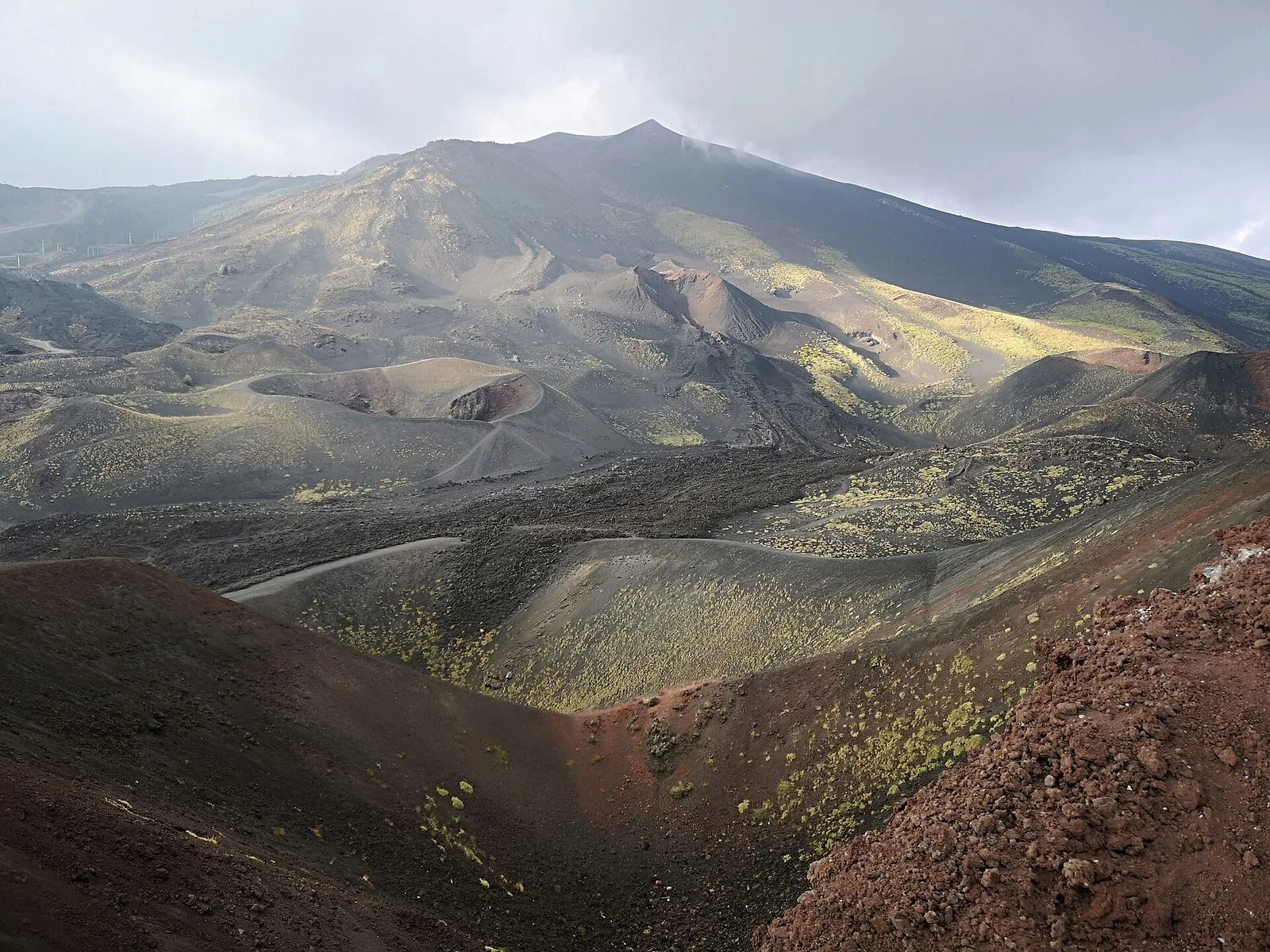 I Crateri Silvestri sul versante sud dell'Etna: coni vulcanici bruni e rossastri con sentieri sul bordo, distese di lava scura punteggiate di arbusti gialli e, sullo sfondo, il pendio del vulcano con la cima fra le nuvole