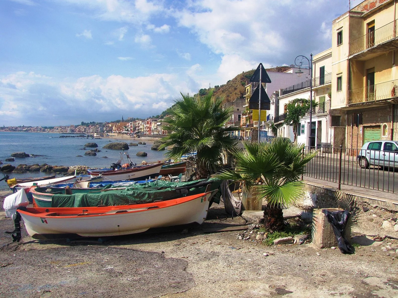 Barche da pesca colorate tirate a secco vicino al mare a Giardini Naxos, con piccole palme, scogli nell'acqua e le case del lungomare che si allungano lungo la baia sotto un cielo con nuvole bianche