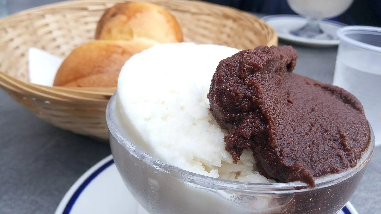 Una coppa di vetro con granita bianca alla mandorla e granita scura al cioccolato, con due brioche dorate in un cestino di vimini sullo sfondo