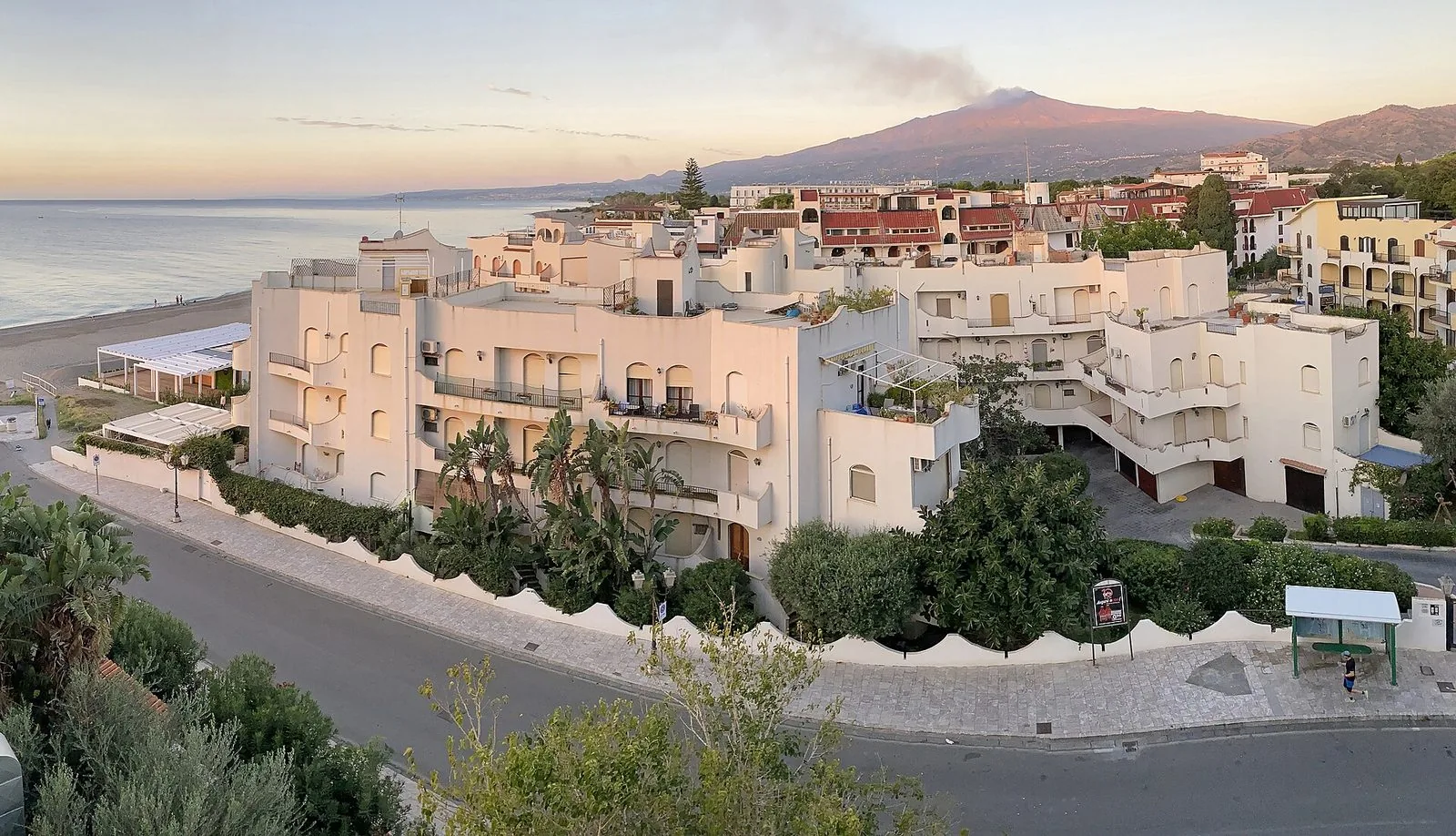 Vista dall'alto di Giardini Naxos al tramonto: edifici bianchi con balconi e giardini, la spiaggia e il mare a sinistra e l'Etna con un pennacchio di fumo sullo sfondo
