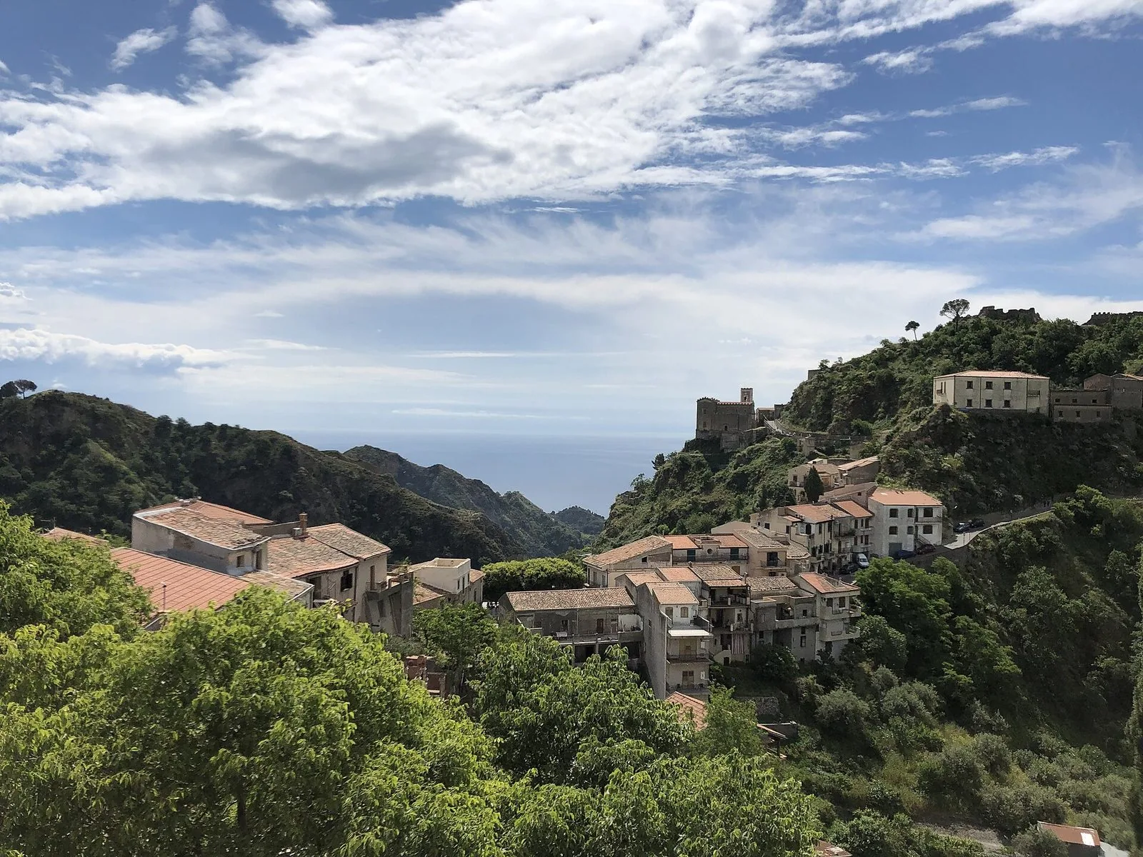 Panorama di Savoca: case con i tetti in coppi fra la vegetazione, la chiesa merlata di San Nicolò su uno sperone roccioso e, in lontananza fra le colline, uno spicchio di mar Ionio sotto un cielo con nuvole leggere