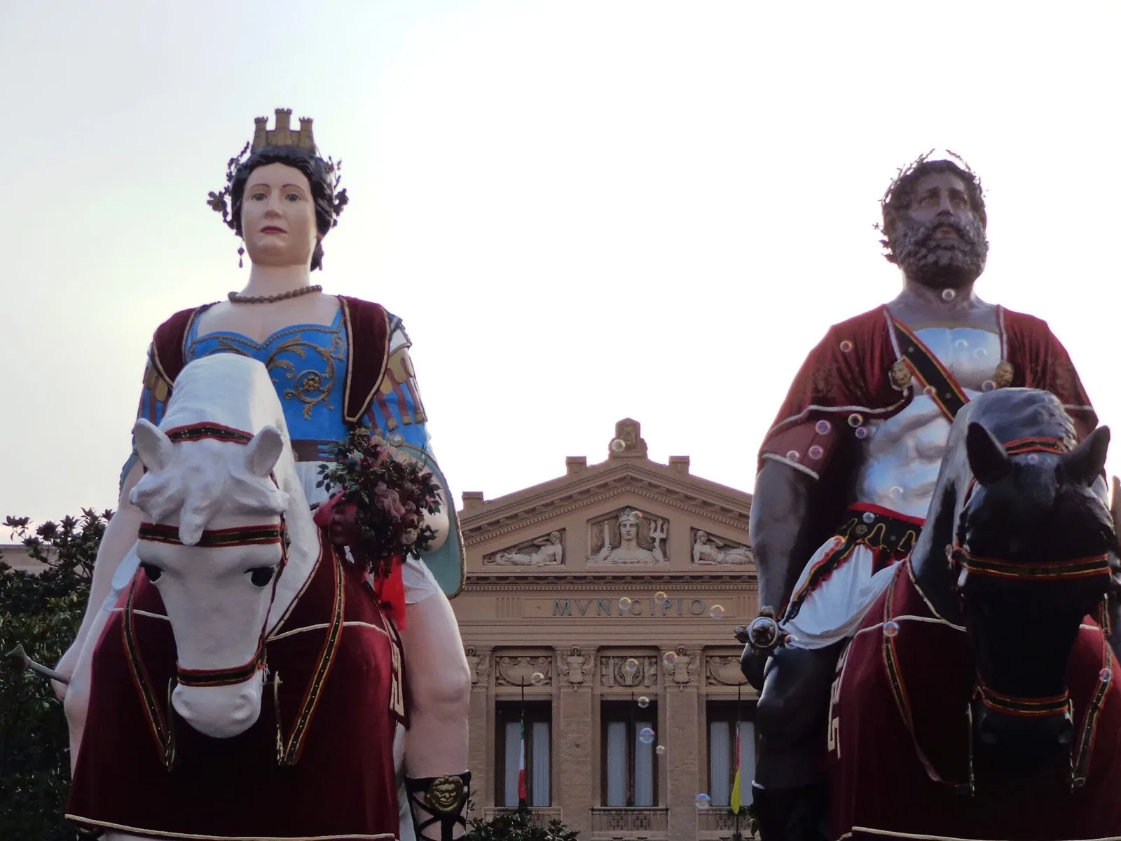 I due Giganti di Messina visti da vicino: a sinistra Mata, con corona turrita e abito azzurro, su un cavallo bianco; a destra Grifone, con barba e corazza argentata, su un cavallo scuro; sullo sfondo la facciata neoclassica del Municipio con la scritta MVNICIPIO